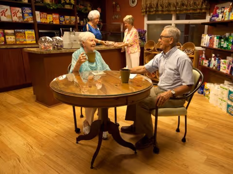 Two elderly people sitting at a round wooden table drinking coffee in a small store or snack area with shelves stocked with cereal, snacks, and household items. In the background, two elderly women are standing near the counter, engaging with each other.