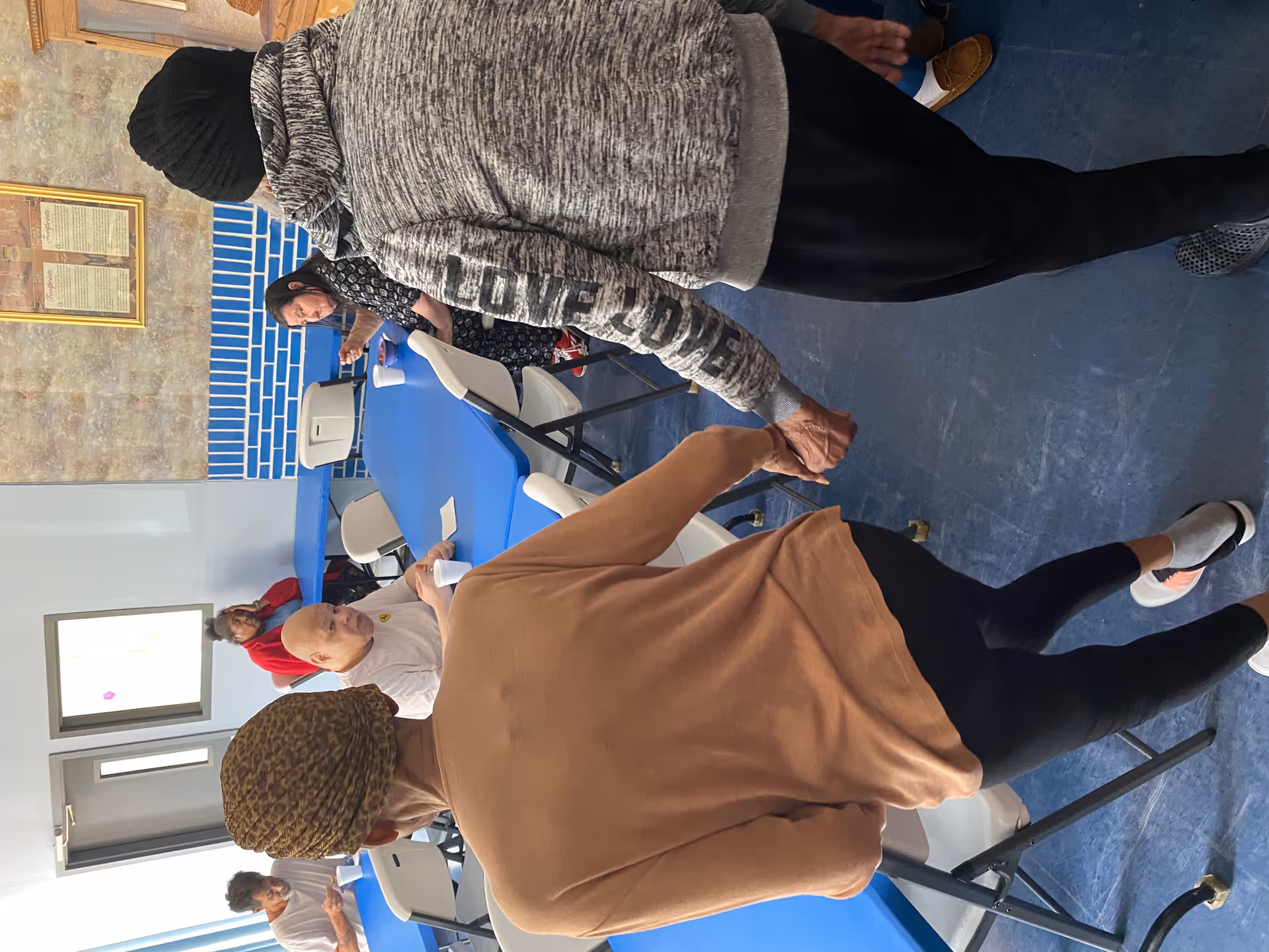 Two elderly people holding hands and walking in a dining area with blue tables and white chairs. Other elderly individuals are seated at the tables, eating and drinking from white cups. The room has a blue and beige wall with framed pictures.