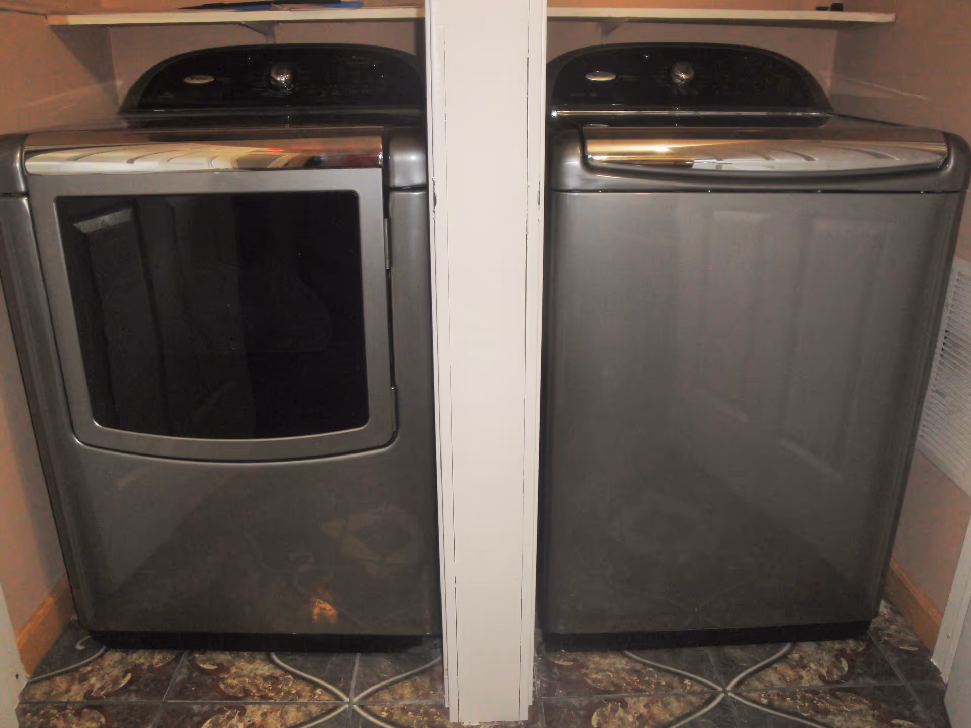 A laundry area with a modern front-loading washing machine on the right and a matching front-loading dryer on the left, separated by a narrow white wall. The floor has patterned tiles and the walls are painted light brown.
