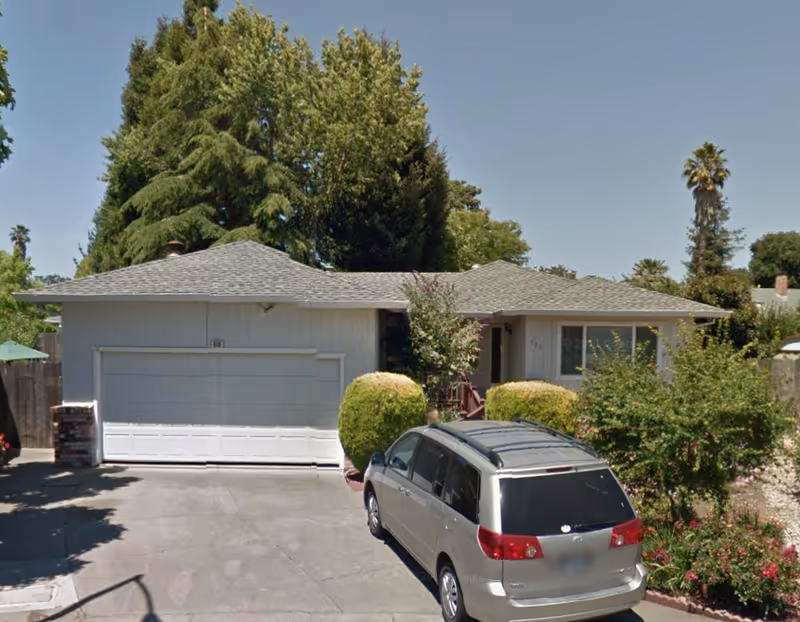 Front exterior view of a single-story house with a two-car garage, a driveway with a silver minivan parked, and surrounding greenery including bushes and tall trees under a clear blue sky.