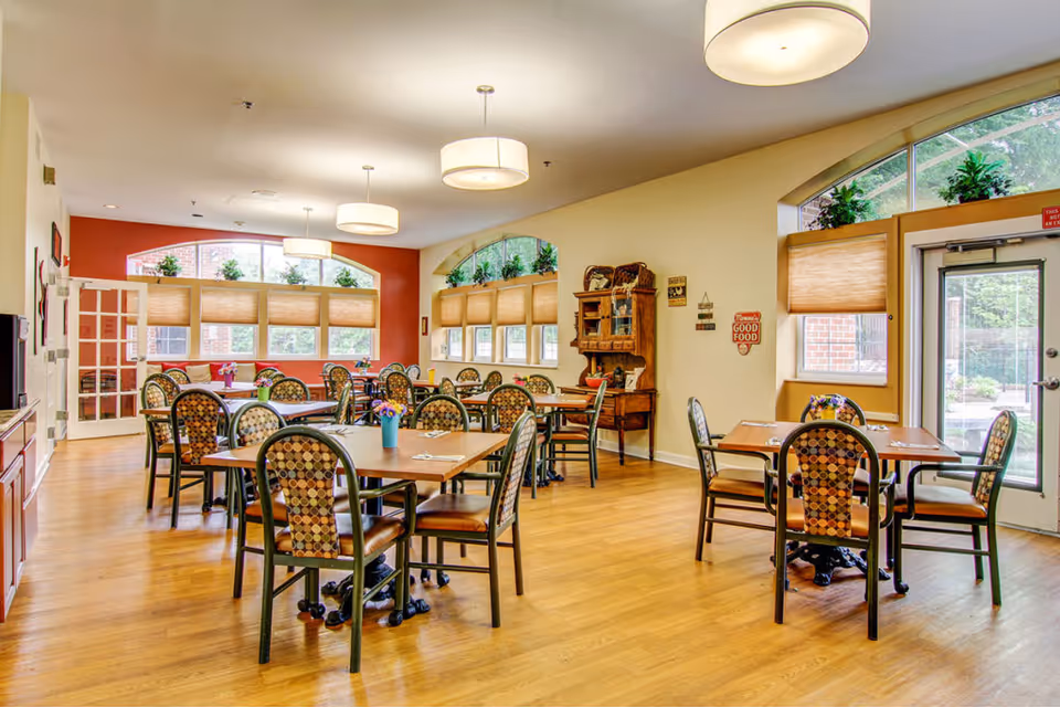 Bright communal dining room with multiple wooden tables and patterned chairs, large arched windows, wood flooring and pendant lights.