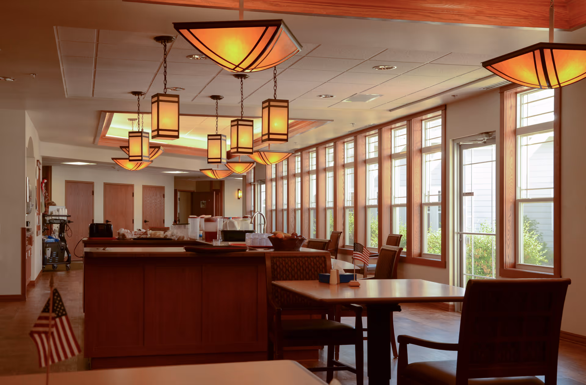 Bright communal dining room with pendant lights, a service counter, tables and large windows along one wall.