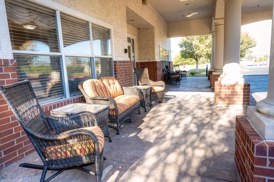Outdoor covered patio area with wicker chairs and a loveseat with striped cushions, a glass-top side table, brick columns, and large windows reflecting the outdoor scenery.