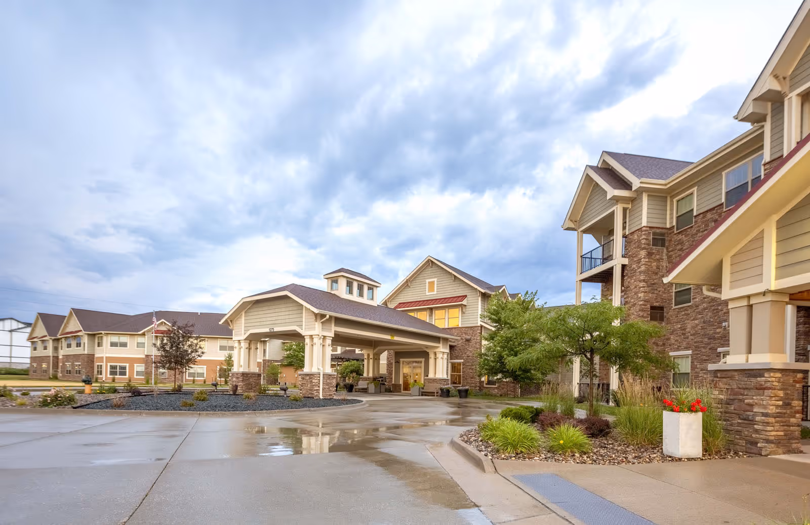 Exterior view of Independence Village of Ankeny showing a large covered entrance with stone and beige siding, surrounded by landscaped greenery and a wet driveway under a cloudy sky.