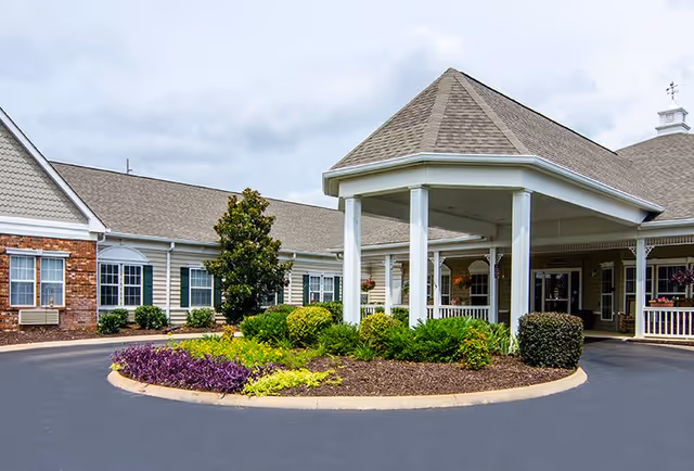 Entrance of a senior living facility with a covered driveway, landscaped garden with shrubs and flowers, and a building with beige siding and brick accents under a cloudy sky.