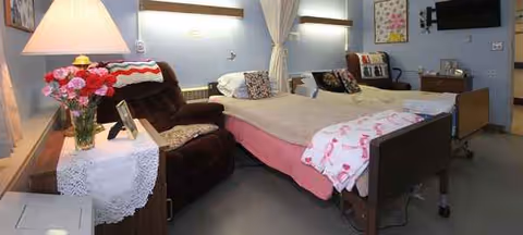 Two beds with blankets, a recliner, flowers, and curtains in a shared bedroom.