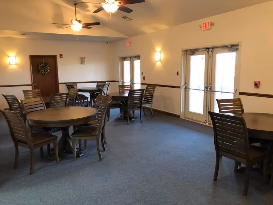 Spacious dining/activity room with round tables and wooden chairs, ceiling fans, and double exit doors.