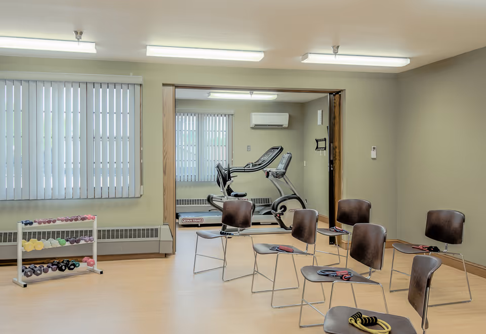 A fitness room with several chairs arranged in a semi-circle, each with resistance bands placed on them. To the left, there is a rack holding various colorful dumbbells. In the background, through an open doorway, a treadmill is visible in an adjacent room with vertical blinds covering the windows.