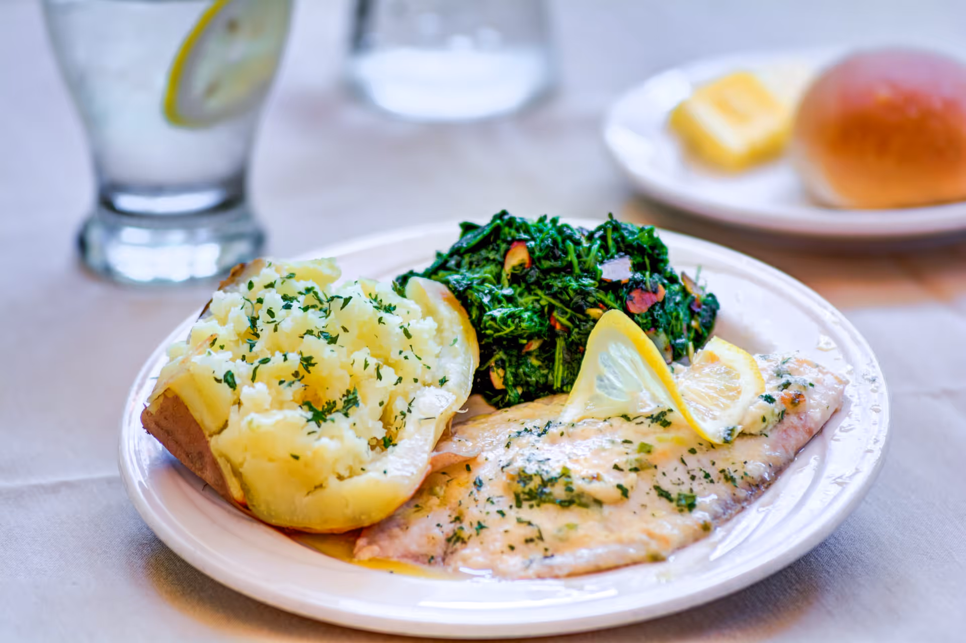 A plate of food featuring a baked potato topped with herbs, sautéed spinach with garlic, and a fillet of fish garnished with lemon slices and herbs. In the background, there is a glass of water with a lemon slice and a plate with a bread roll and a pat of butter.