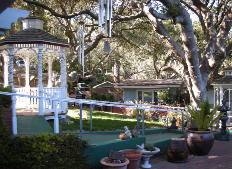 Outdoor garden area with a white gazebo, large trees, potted plants, wind chimes, and a green ramp with metal handrails leading up to the gazebo. There is a small building in the background surrounded by greenery.