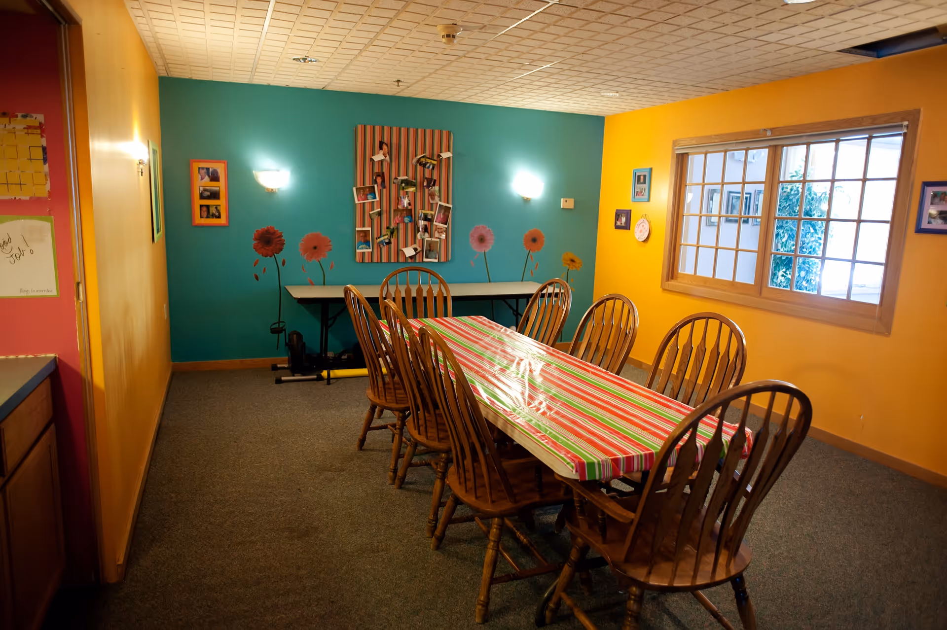 Bright communal dining room with a long striped-tablecloth table surrounded by wooden chairs and colorful painted walls.