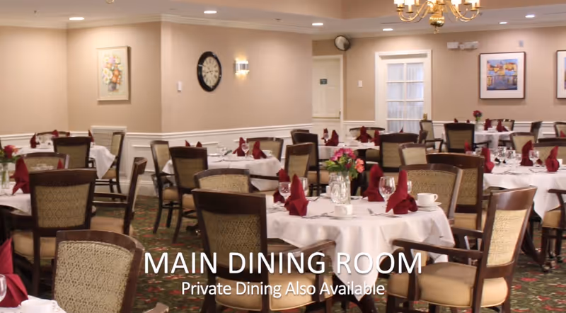A spacious dining room with multiple round tables covered in white tablecloths, each set with folded burgundy napkins, glassware, and floral centerpieces. The room has beige walls adorned with framed artwork and a clock, with cushioned wooden chairs arranged around the tables. A chandelier hangs from the ceiling, providing warm lighting.