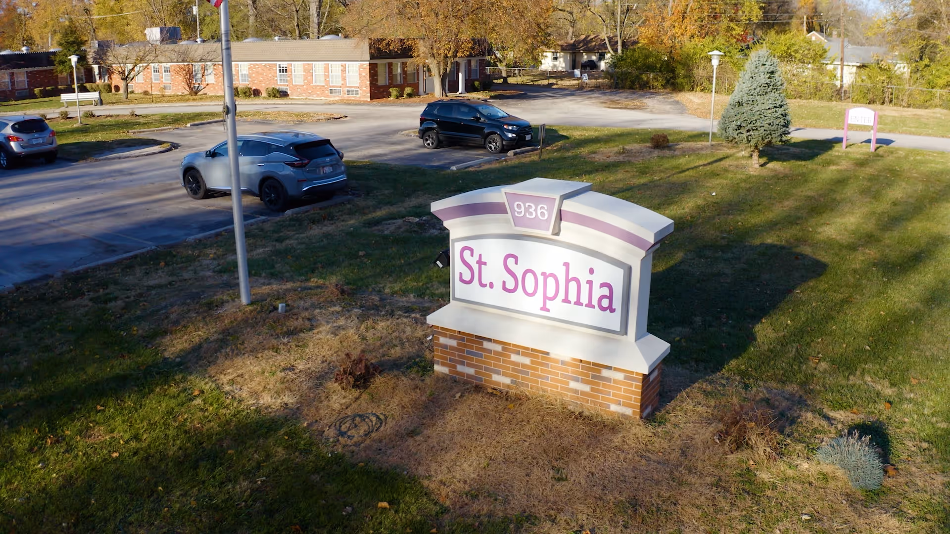 Outdoor view of the St. Sophia Health & Rehabilitation Center sign with the number 936 on top, situated on a grassy area near a parking lot with several parked cars and a brick building in the background.