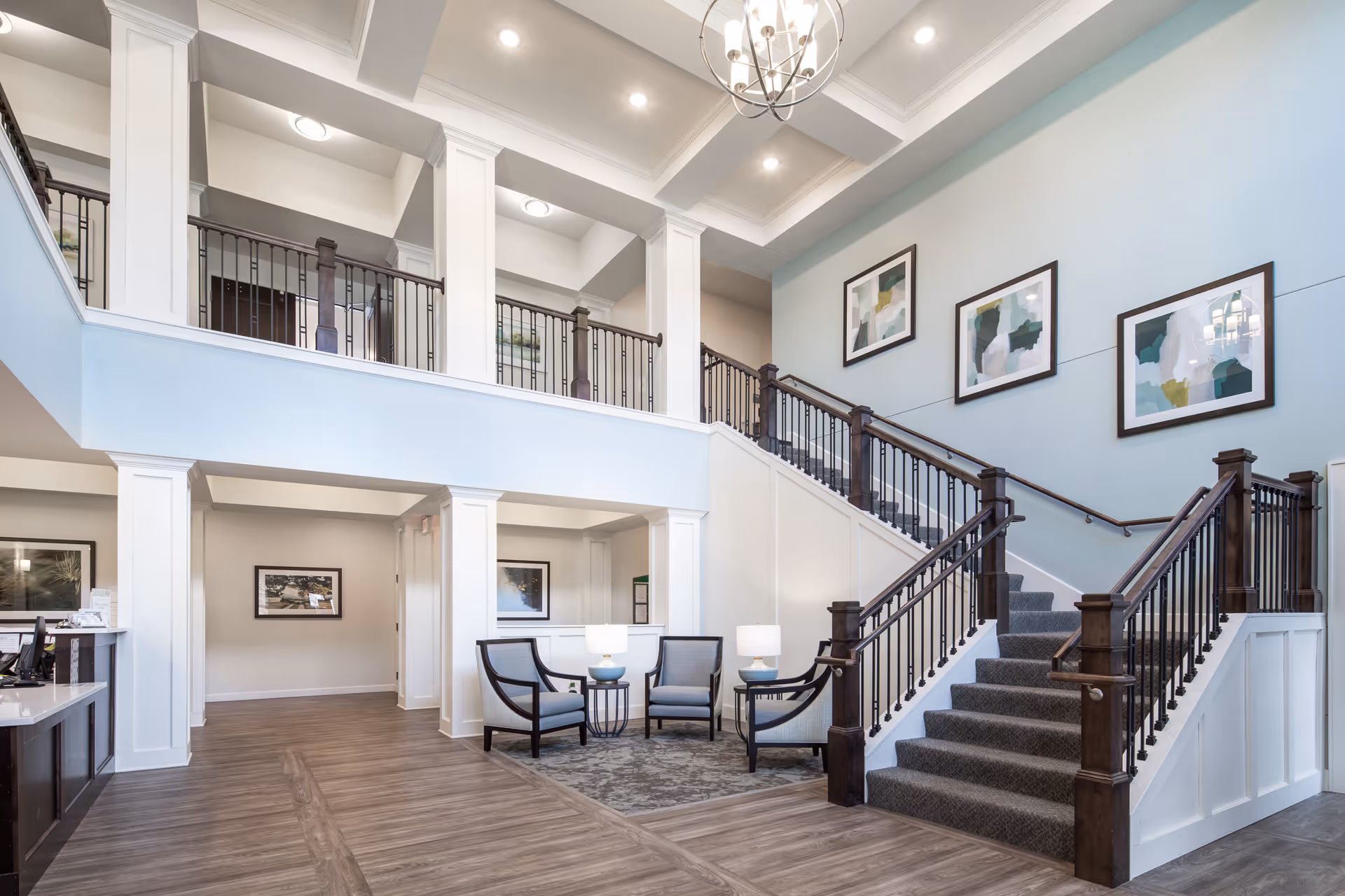 Bright and spacious interior of a senior living facility featuring a carpeted staircase with dark wooden railings, light blue walls adorned with abstract artwork, a seating area with four chairs and two small tables with lamps, and a reception desk on the left side.