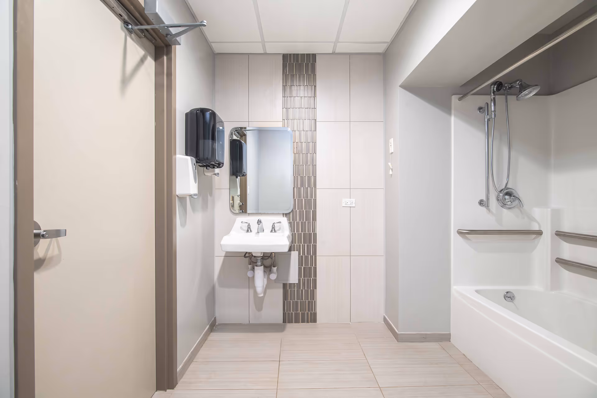 A clean and modern bathroom featuring a white bathtub with a showerhead and grab bars, a wall-mounted sink with a mirror above it, and a soap dispenser and paper towel holder mounted on the wall. The walls are tiled with a vertical strip of decorative tiles behind the sink, and the floor is covered with light-colored tiles.