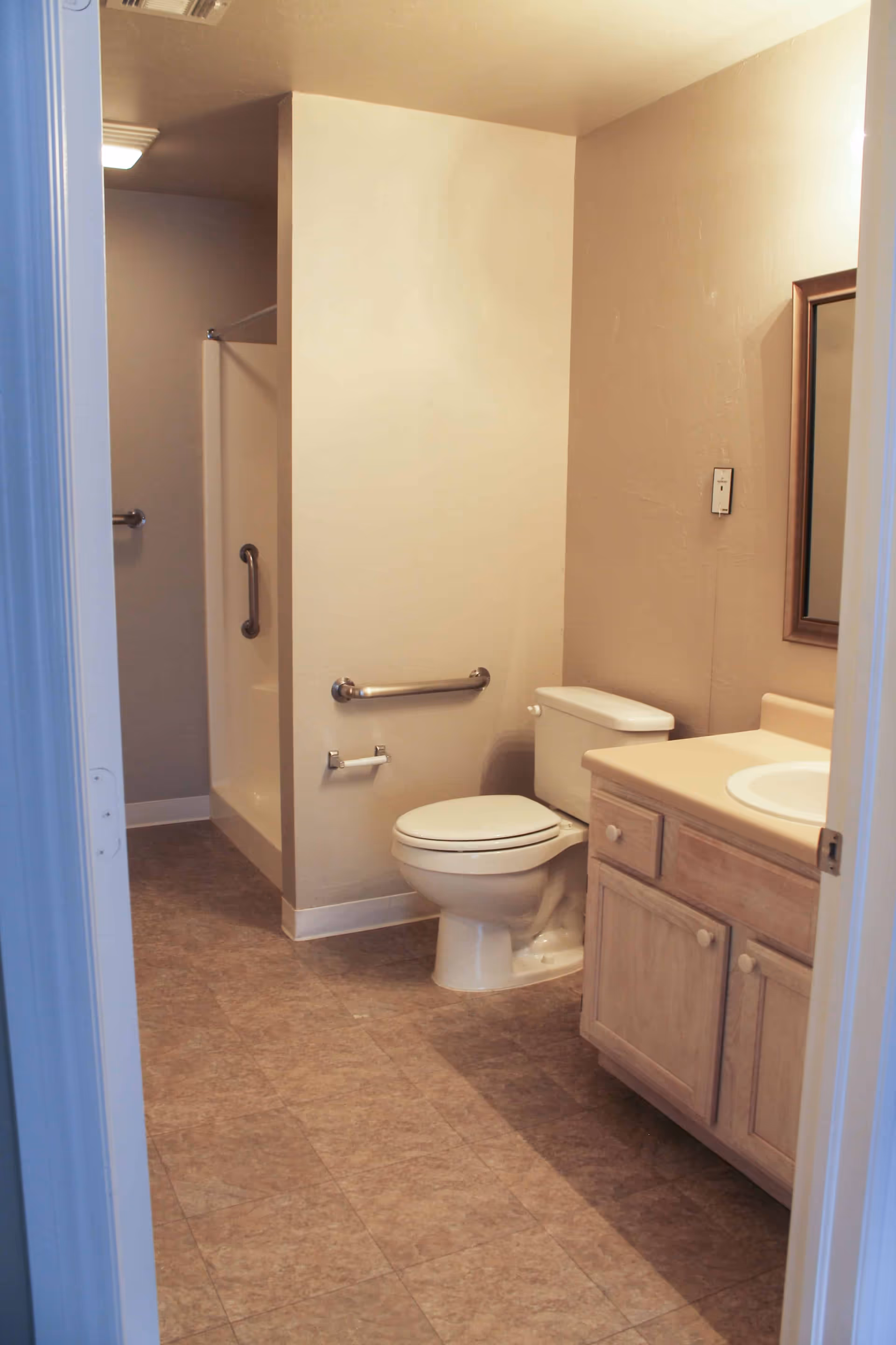 A bathroom with a toilet, a sink with a wooden cabinet, a mirror, and a walk-in shower with grab bars. The walls are beige and the floor has brown tiles.