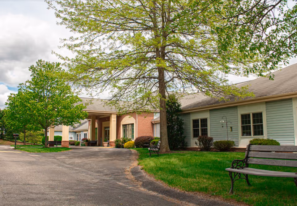 Exterior view of St. Barnabas - Arbors III facility showing a driveway leading to the entrance with a covered porch. The building has a mix of brick and light blue siding with several windows. There are green trees and bushes around the building, along with benches on the grass near the driveway.