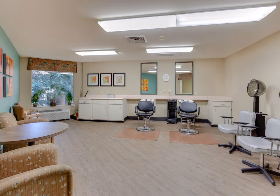 Bright salon-style common room with barber chairs and mirrors at a counter, a seating area with tables, and a window with plants.