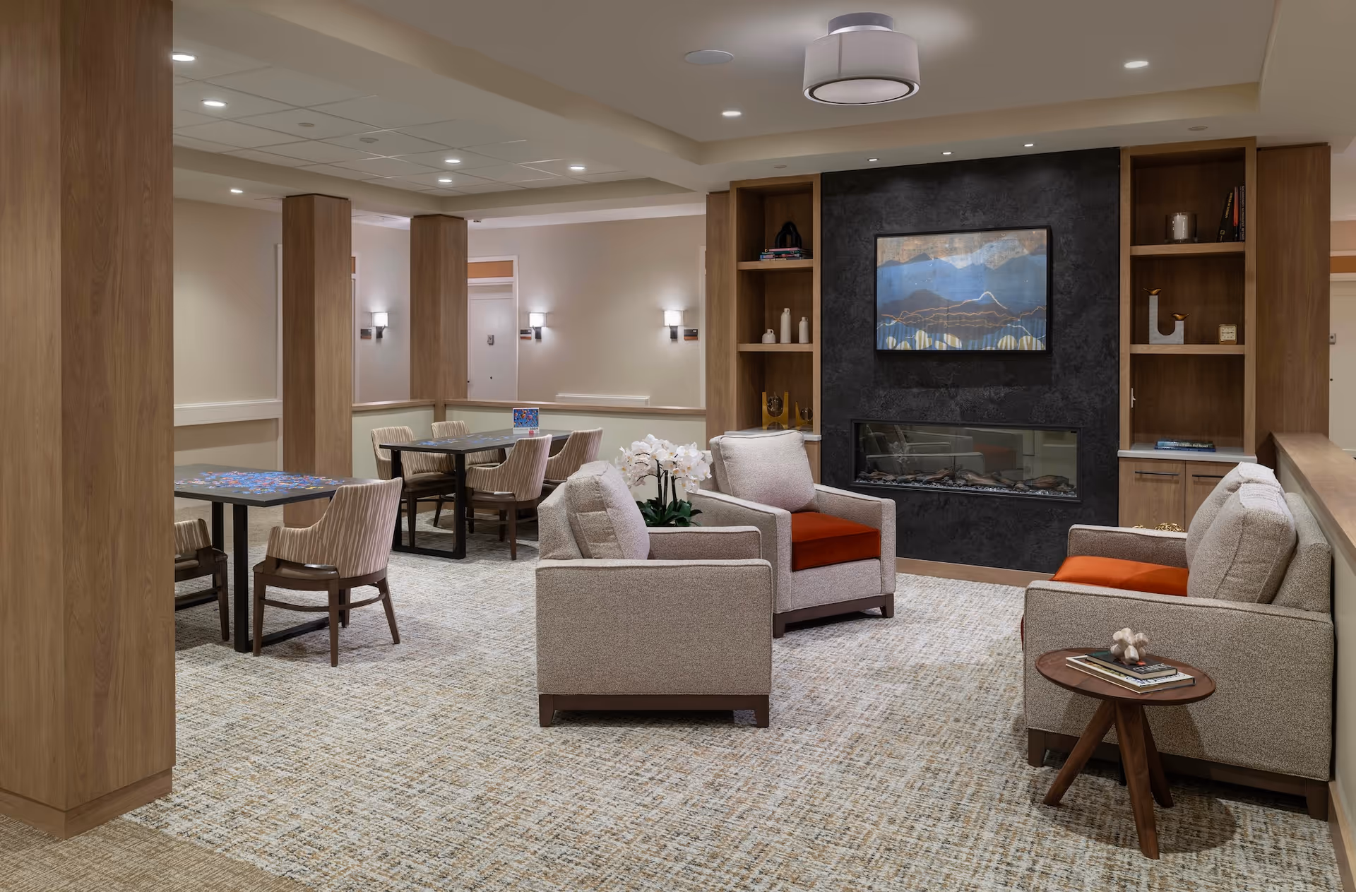 A cozy living room area in Monarch Coopers Corner featuring beige armchairs and a sofa with orange cushions arranged around a modern fireplace set in a dark textured wall. Built-in wooden shelves flank the fireplace, decorated with books and ornaments. In the background, there are tables and chairs for games or dining, with soft lighting and neutral-colored walls and carpet.