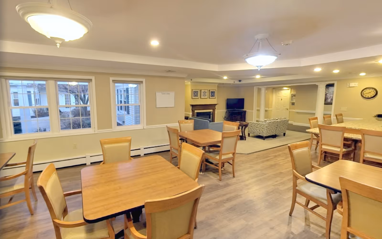 A spacious common area in a senior living facility featuring multiple wooden tables with chairs arranged around them. The room has large windows letting in natural light, a seating area with a TV and fireplace, and warm overhead lighting. The flooring is wood-style, and the walls are painted in a neutral beige tone.