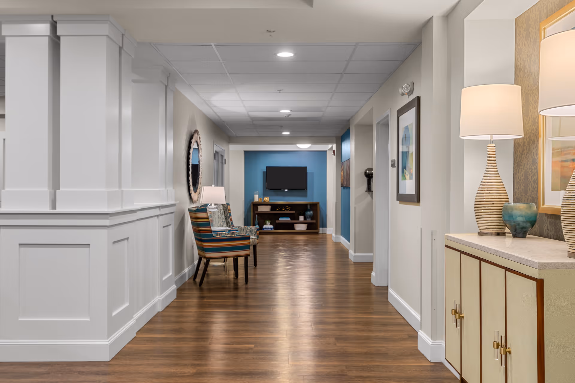 Well-lit interior corridor lounge with wood floors, seating, decorative lamps, and a TV at the far end.