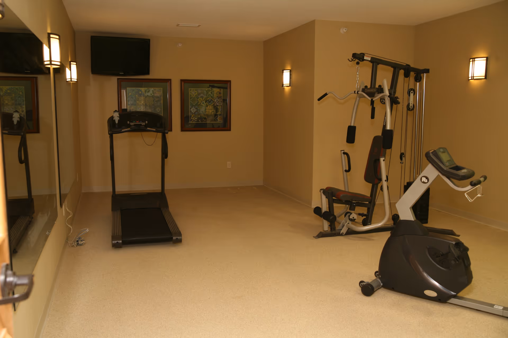 A small fitness room with exercise equipment including a treadmill, a multi-function weight machine, and a stationary bike. The room has beige walls, two framed pictures on the wall, a wall-mounted TV, and wall sconces providing lighting. A large mirror is visible on the left side.