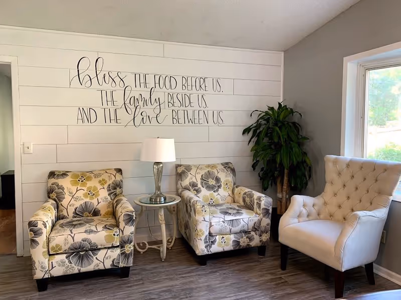 A cozy seating area with two floral patterned armchairs and one beige tufted armchair arranged around a small round side table with a lamp. A large green plant is placed in the corner near a window letting in natural light. On the wall behind the chairs is a decorative sign that reads, 'Bless the food before us, the family beside us, and the love between us.'
