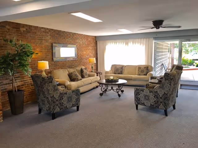 A cozy living room with two beige sofas and two patterned armchairs arranged around a round coffee table. The room features a brick accent wall with a rectangular mirror and two table lamps. Large windows with sheer curtains allow natural light to brighten the space, and a ceiling fan is mounted above. A sliding glass door leads to an outdoor patio area.