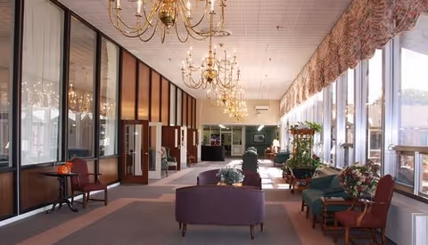 Long sunlit senior living lounge with sofas and chairs arranged along a carpeted corridor, chandeliers overhead and large windows on one side.