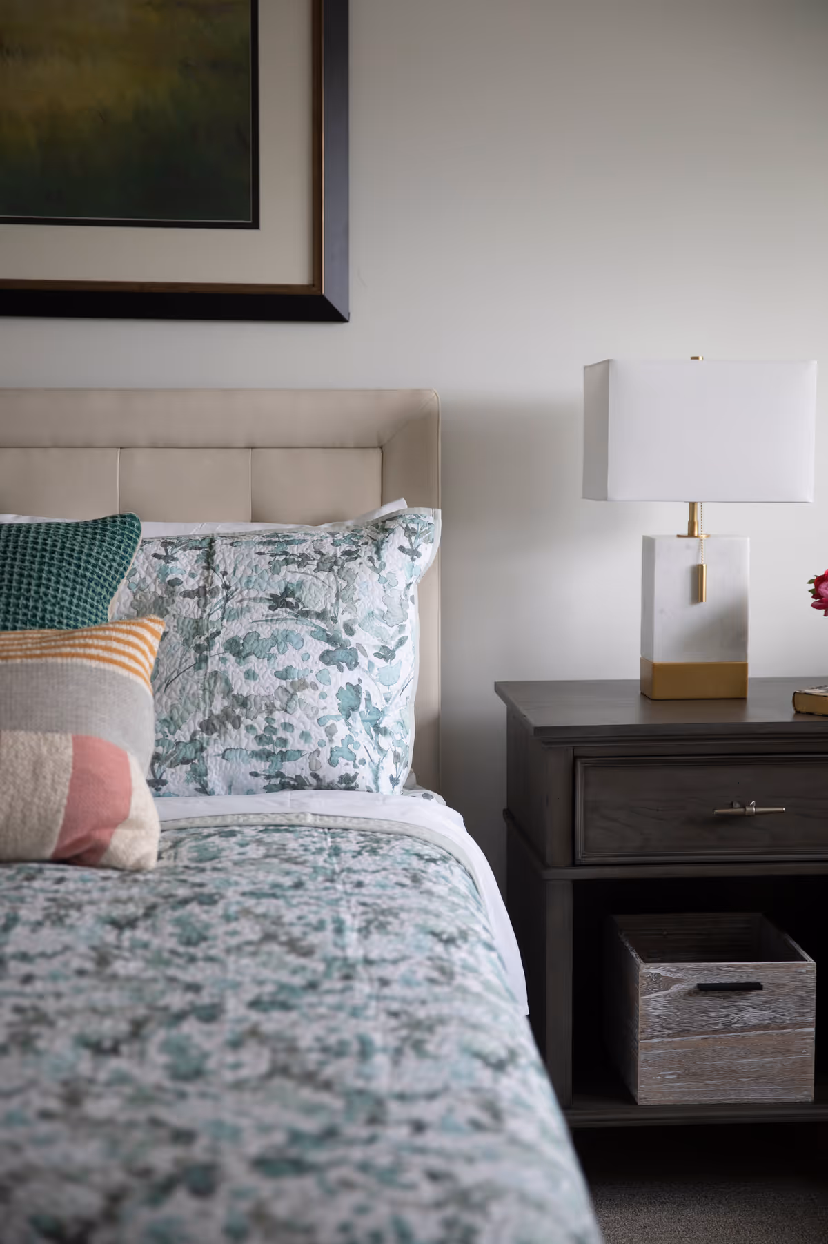 A neatly made bed with green-and-white patterned bedding, an upholstered headboard, and a dark wooden nightstand holding a white-and-gold lamp.