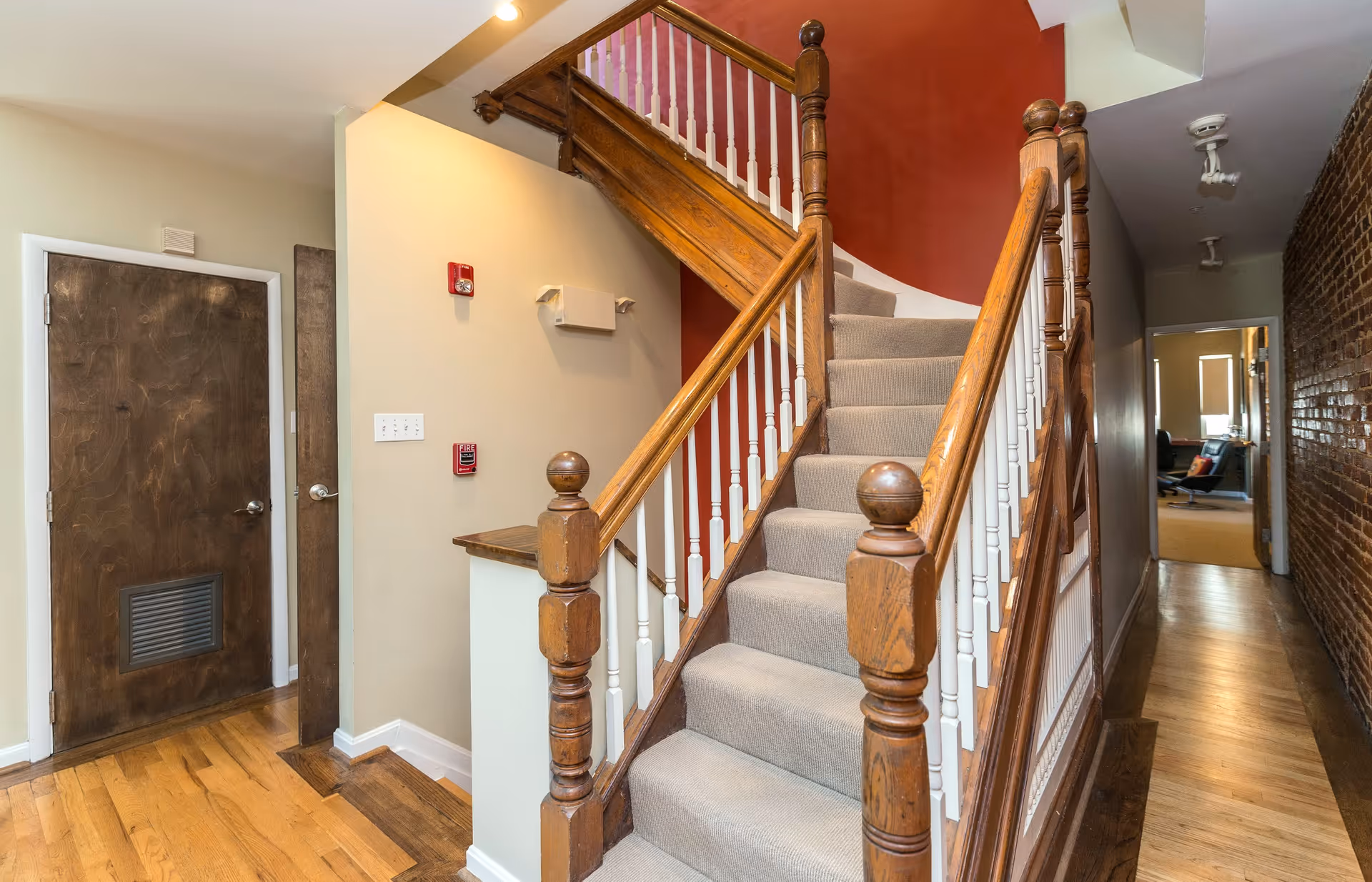 Interior view of a hallway featuring a wooden staircase with beige carpet and white spindles. The walls are painted beige and red, with a wooden door on the left and a long corridor on the right leading to a room with a window and furniture.