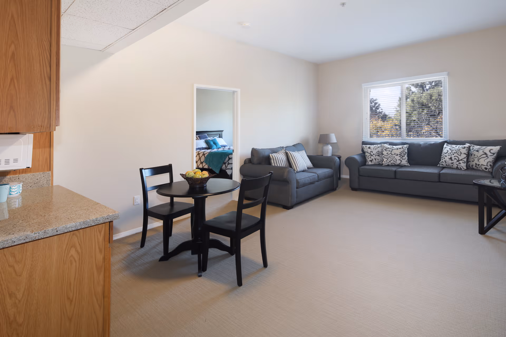 Bright furnished living room with two gray sofas, a small round dining table with chairs, a window, and a partial view into a bedroom and kitchen area.