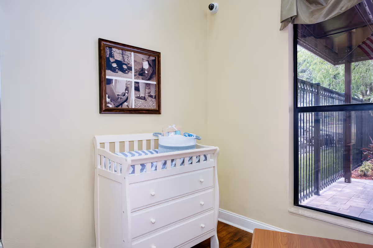A small corner of a room with a white changing table that has three drawers and a striped changing pad on top. Above the changing table is a framed picture showing four close-up images of feet wearing different shoes. To the right, there is a window with a view of an outdoor patio area with a black metal fence and greenery.