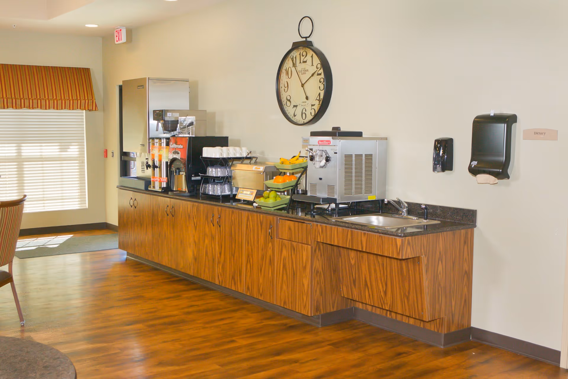 A beverage and snack station in a senior living facility with a coffee machine, juice dispensers, cups, fruit baskets, a sink, and a large wall clock above. The area has wooden cabinets and a hardwood floor, with a window covered by a striped valance in the background.