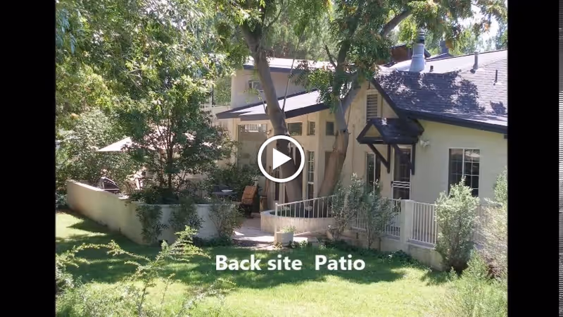 A backyard patio area of a residential-style building surrounded by trees and greenery. The patio is enclosed by a low wall and has outdoor seating. The building has light-colored walls and a dark roof with a small awning over the door. The text 'Back site Patio' is visible on the grass in front of the patio.