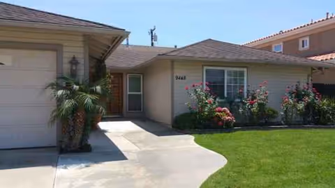 A single-story beige house with a garage, walkway, and blooming shrubs in the front yard.