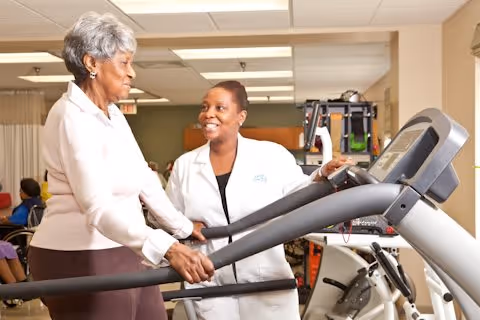 An elderly woman using a treadmill in a healthcare facility while being assisted and encouraged by a female healthcare professional wearing a white coat.