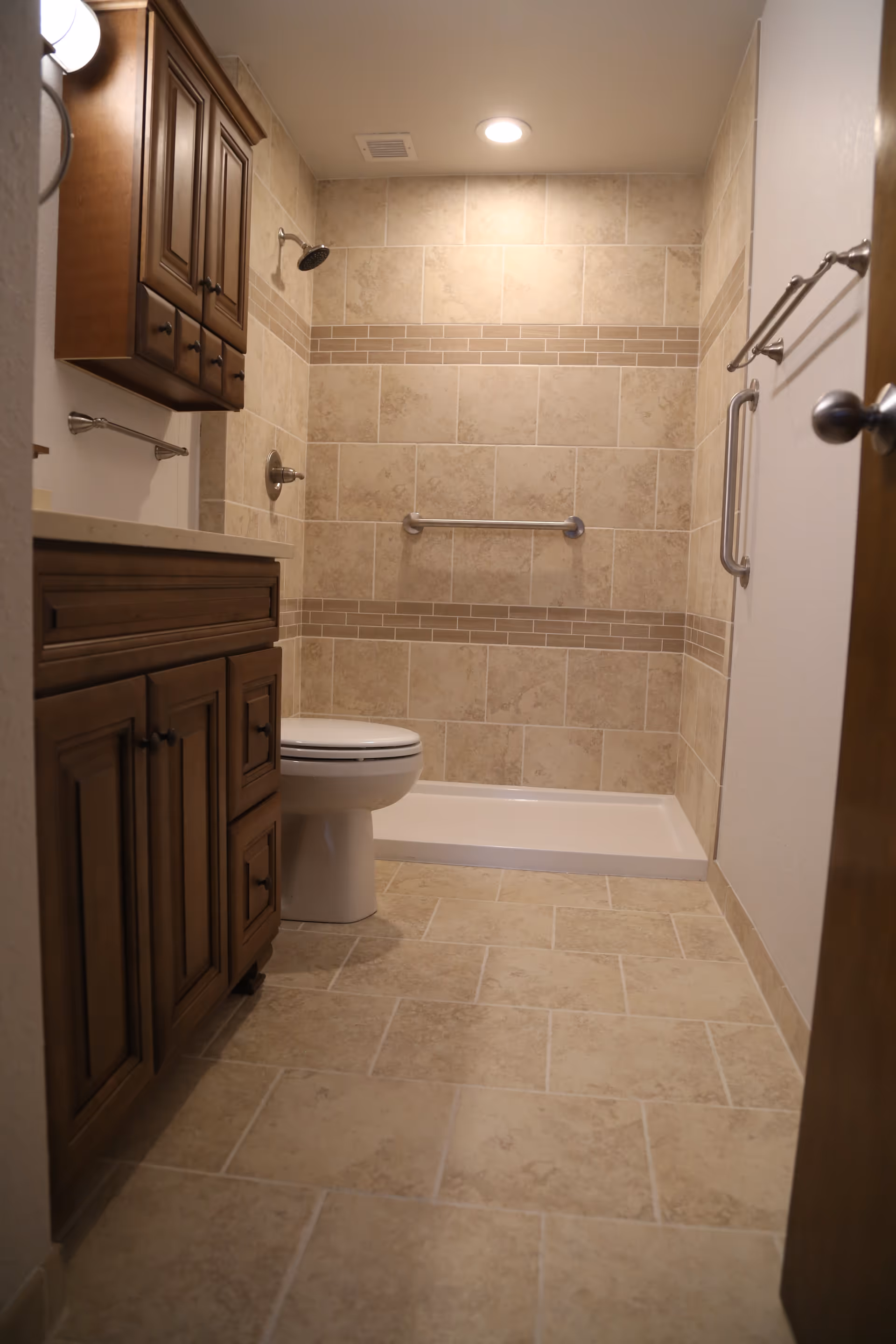 Tiled bathroom with a walk-in shower, grab bars, toilet, and wooden vanity cabinets.