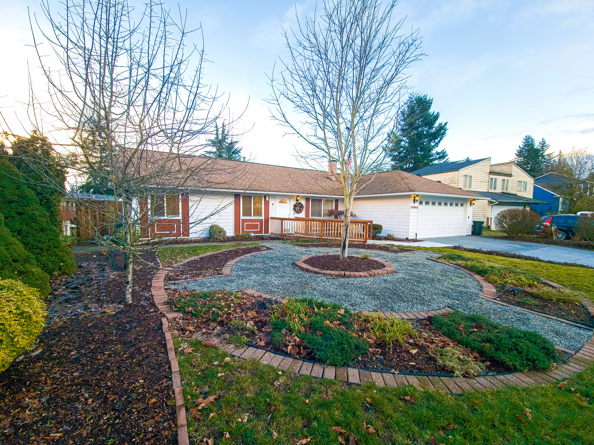 Front exterior view of a single-story residential building with a brown roof and white siding, featuring a wooden ramp leading to the front door decorated with a wreath. The yard has a circular gravel area with a tree in the center, surrounded by landscaped plants and bushes. Neighboring houses and a driveway with parked cars are visible.
