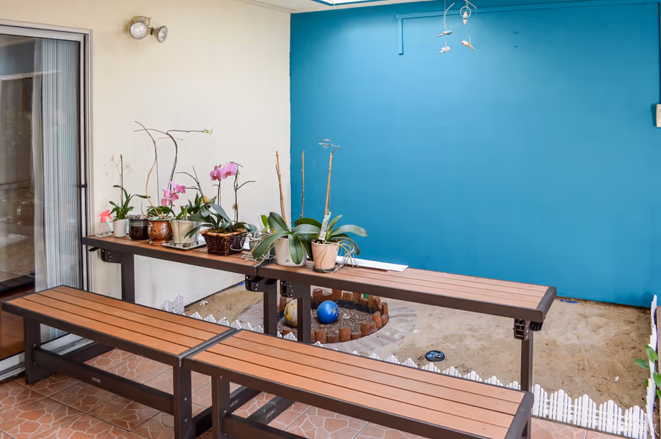 Outdoor patio area with two wooden benches and a long wooden table holding several potted plants, including orchids. The background features a blue wall and a small sandy garden area bordered by a white picket fence. A sliding glass door is visible on the left side.