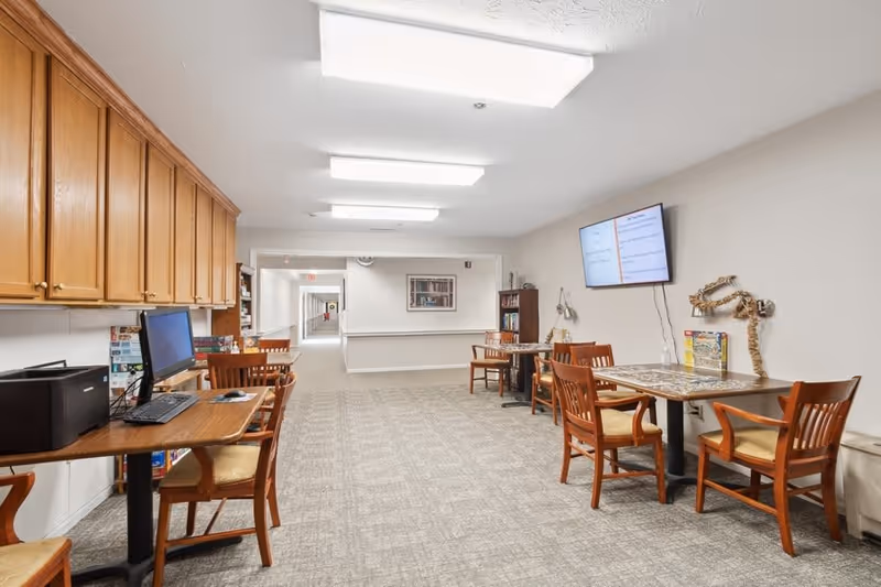 A well-lit interior common area with carpeted flooring, wooden chairs, and tables. On the left side, there is a desk with a computer and printer. On the right side, two tables with chairs are positioned against the wall, one with a mounted TV screen above it. The walls are painted light beige, and there are overhead fluorescent lights. A hallway extends from the back of the room.