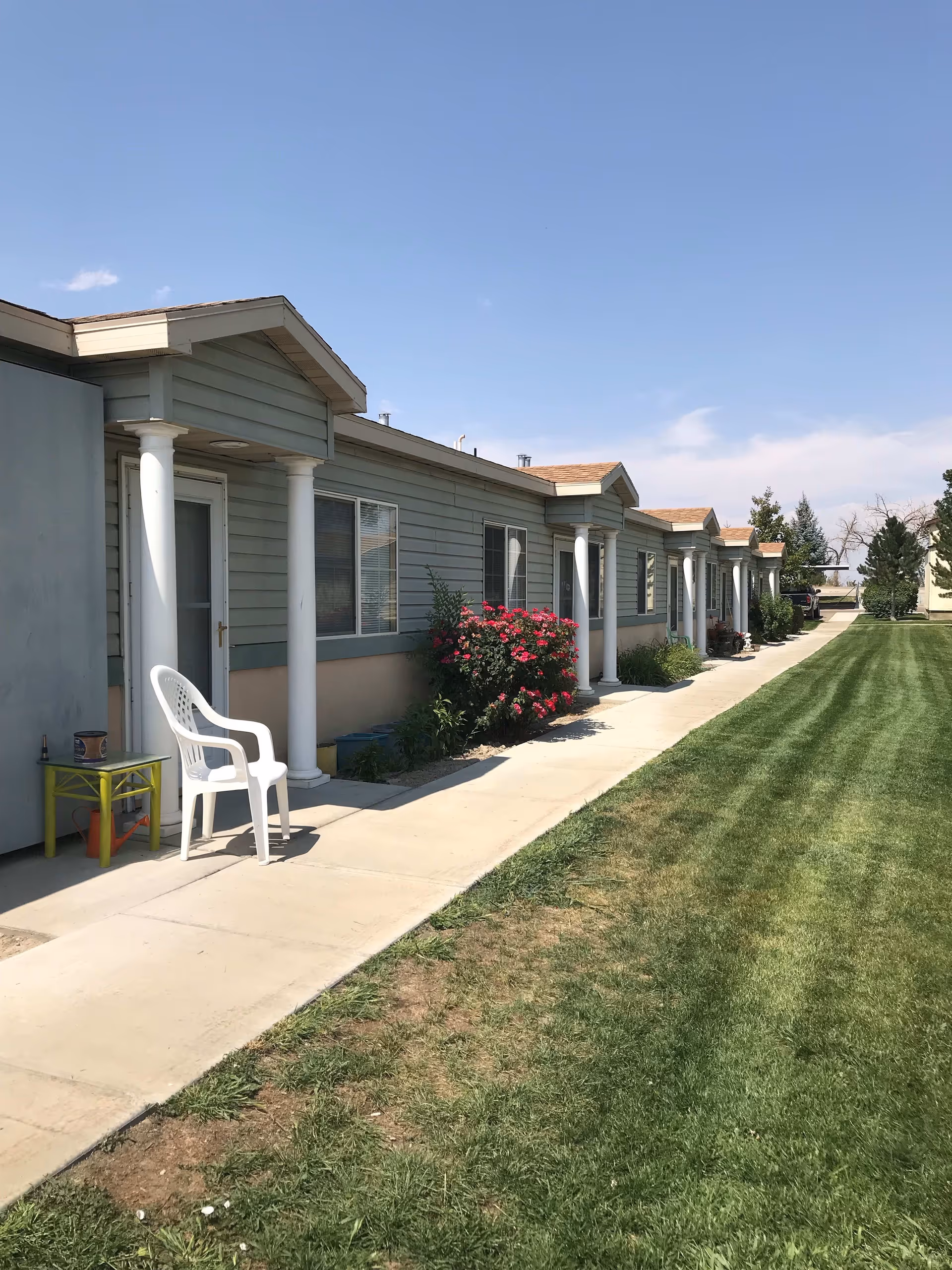 Single-story senior living building exterior showing a row of entrances with white columns, a sidewalk, lawn and a white plastic chair.
