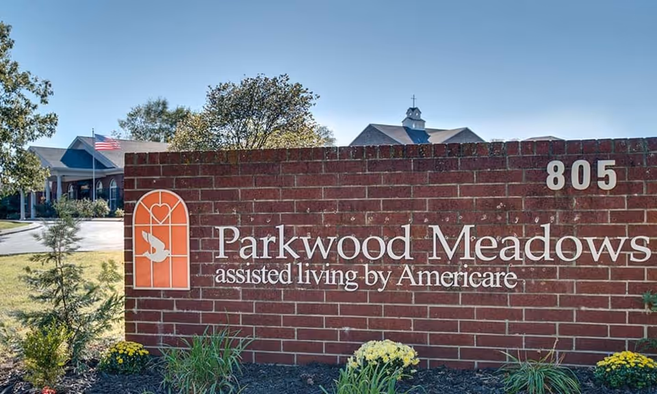 A brick sign for Parkwood Meadows assisted living by Americare with the number 805 on it, surrounded by plants and flowers. In the background, there is a building with a flagpole flying the American flag and trees under a clear blue sky.