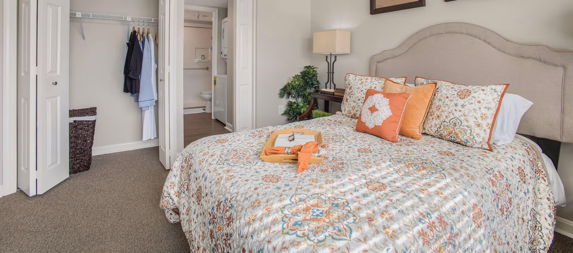 A bedroom with a bed covered in a patterned quilt and several decorative pillows. A wooden tray with a book, glasses, and a cloth is on the bed. To the left, there is an open closet with hanging clothes and a laundry basket. In the background, a bathroom with a toilet and a stacked washer and dryer is visible. A small table with a lamp and a plant is beside the bed.