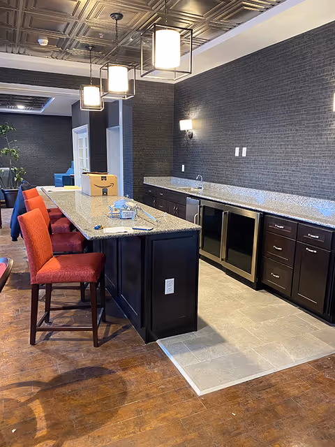 Communal kitchen/bar area with a long granite island, red bar stools, pendant lights, dark cabinets, sink and under-counter refrigerators.