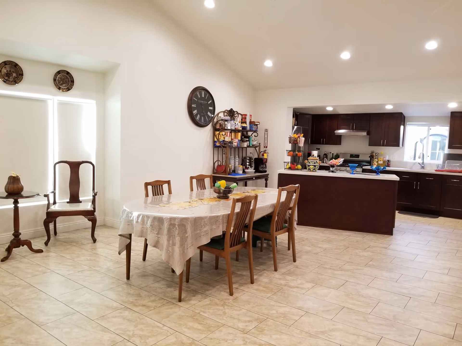 A bright and spacious dining area with a rectangular table covered with a lace tablecloth and six wooden chairs. To the left, there is a wooden armchair and a small round side table with a decorative item. The back wall features a large clock and a metal rack filled with various food items and kitchen supplies. The kitchen area in the background has dark wooden cabinets, a stove with a range hood, a sink under a window, and a kitchen island with decorative bowls and fruit baskets on top. The floor is tiled with light-colored tiles, and recessed ceiling lights illuminate the space.