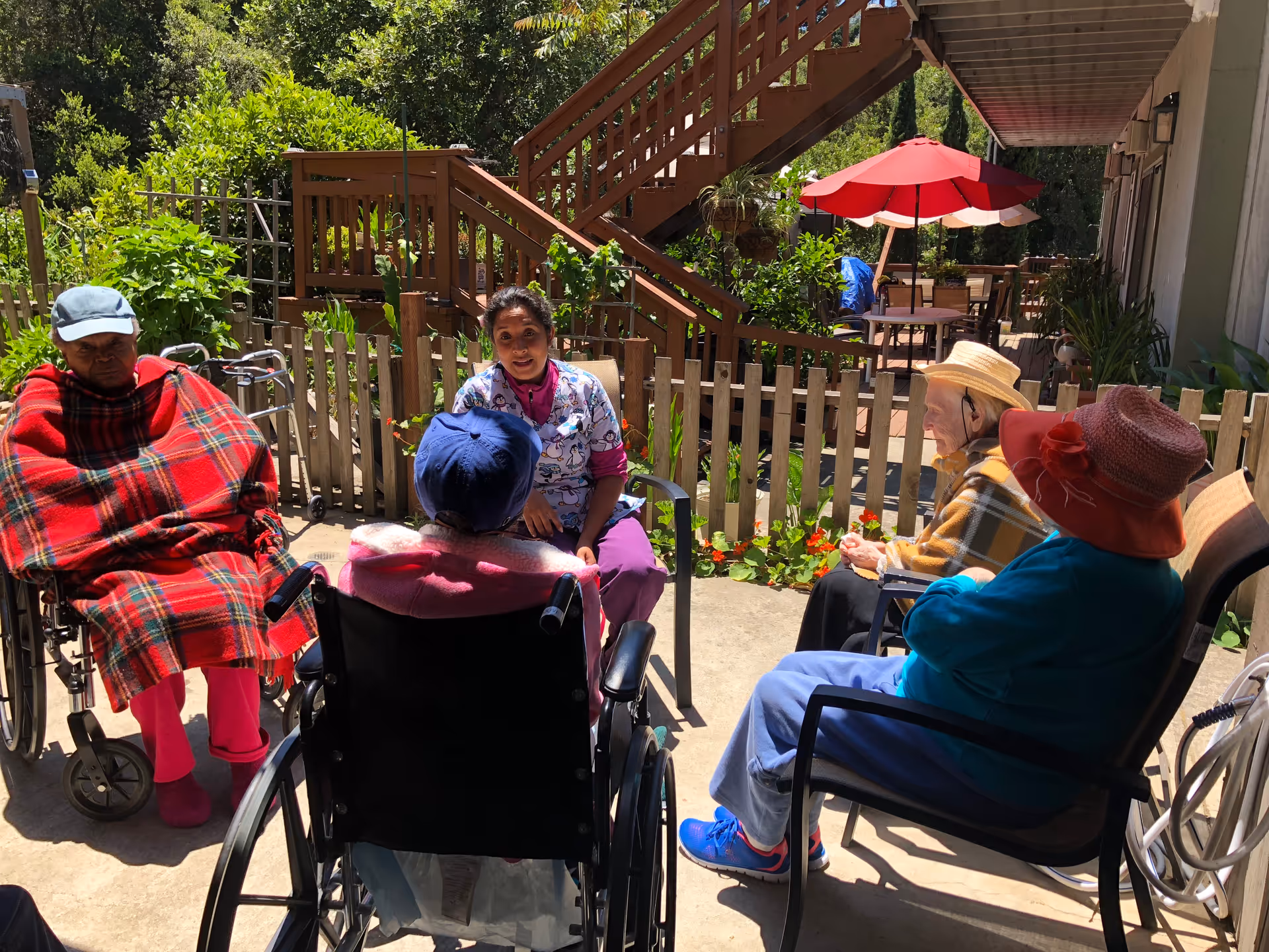 A caregiver and four elderly residents, some in wheelchairs, sitting together in a sunny outdoor courtyard with a wooden staircase, picket fence and red patio umbrella.