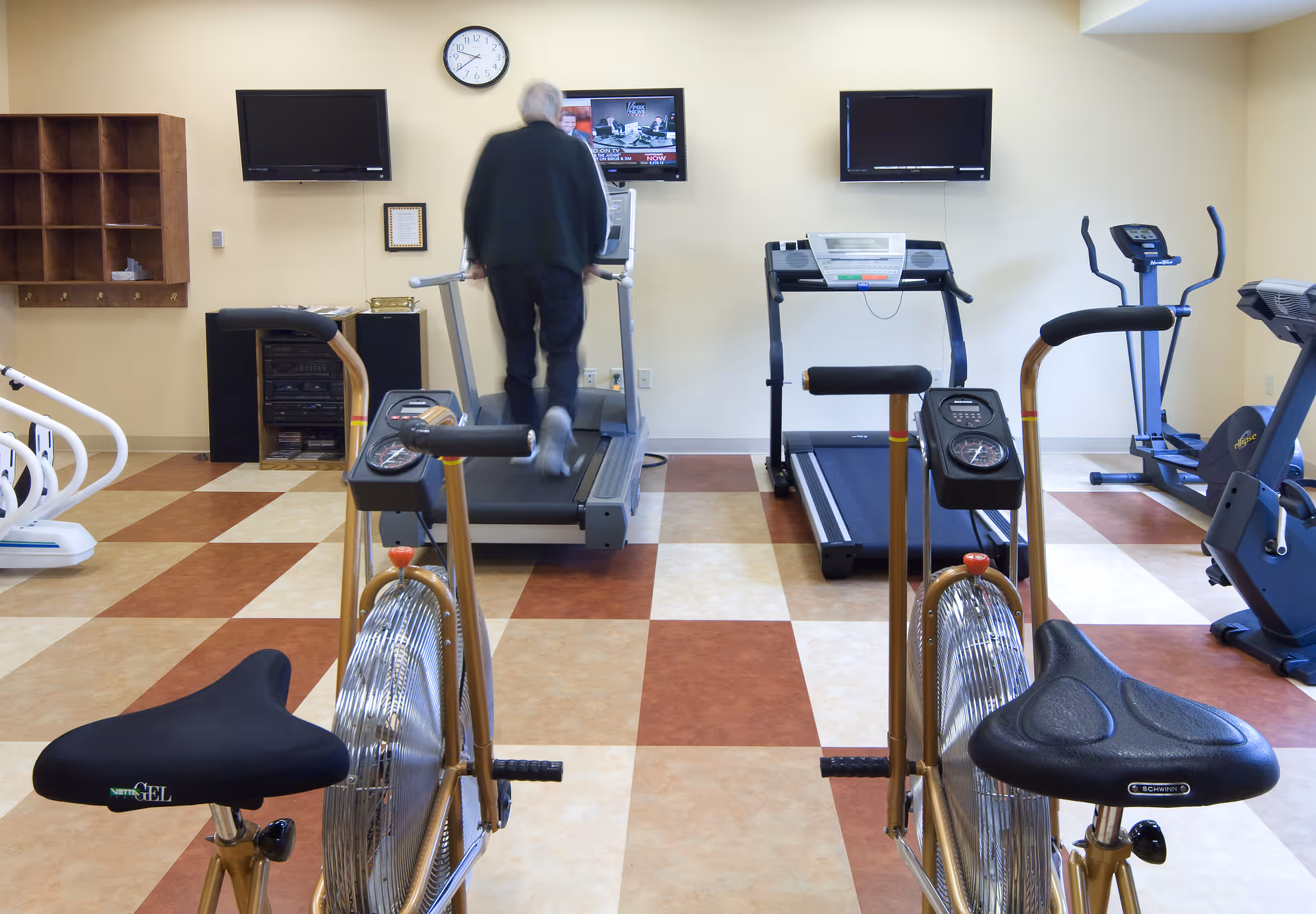 An elderly person walking on a treadmill in a fitness room with exercise bikes, treadmills, and other cardio equipment. The room has a checkered floor pattern and three wall-mounted televisions.