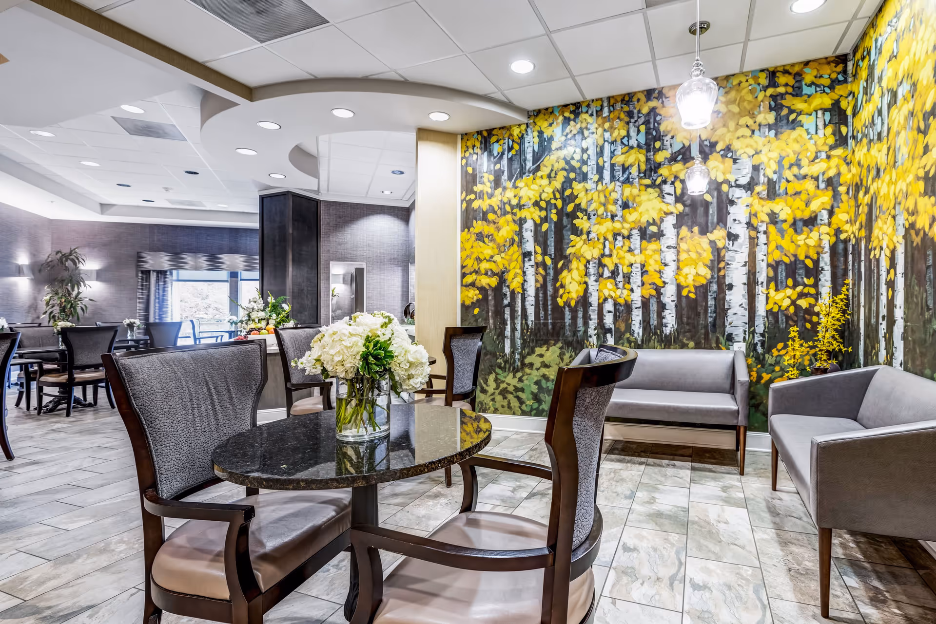 A bright and modern common area in a senior living facility featuring a round black marble table with a vase of white flowers, surrounded by four cushioned chairs. In the background, there are two gray sofas against a wall with a large mural of birch trees with yellow leaves. The room has tiled flooring and recessed ceiling lights.
