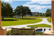 View of a green outdoor area with a well-maintained lawn, trees, a curved walking path, and a small water fountain, seen from a shaded porch with a wooden bench in the foreground.